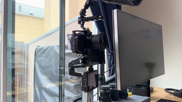 Sony A7 IV in a cage on a cheese plate, mounted to an Ergotron monitor arm above the desk, with the dummy battery adapter hanging below and lenses on the desk
